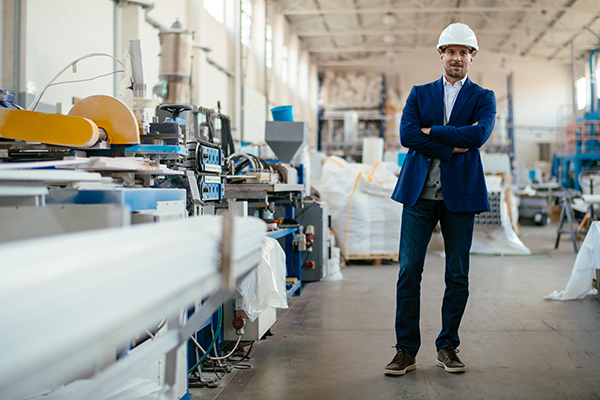 Geschäftsmann mit einem Baustellenhelm der in einer Industriehalle steht neben Maschinen